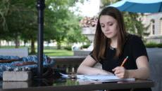 Student sits at a table outside making notes on a paper.